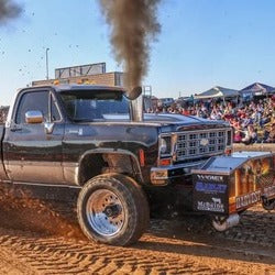 Black pickup truck with a large trailer on a dirt track, surrounded by spectators and dust.